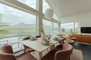 A modern dining area with a view of a lake and mountains, furnished with brown chairs and a large table set for a meal.