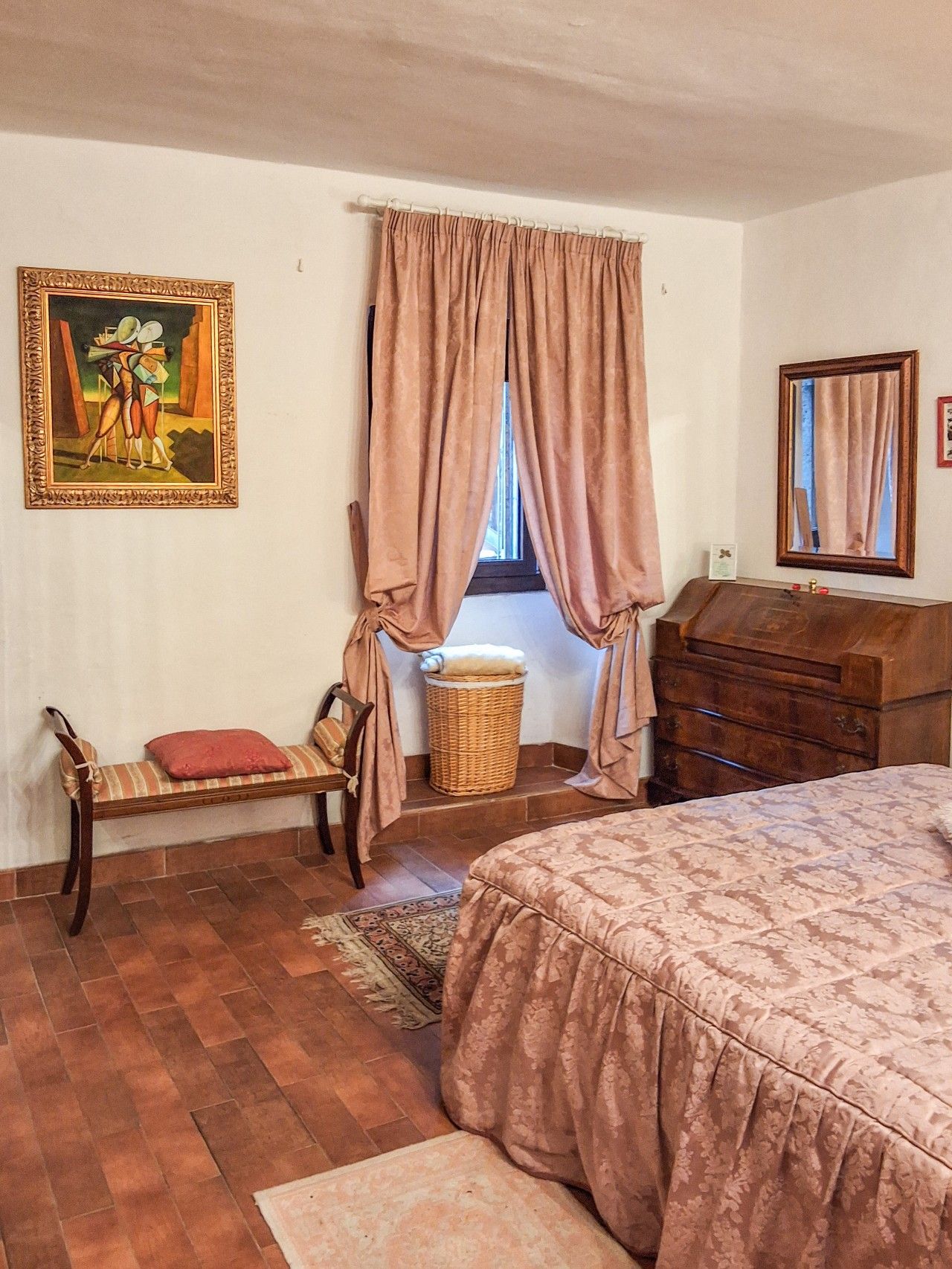 A bedroom with a bed, a wooden dresser, a chair, a wicker basket, a painting, and a window with curtains.