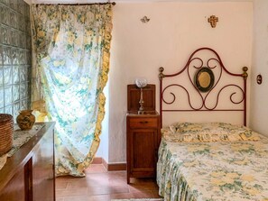 Un dormitori amb un llit, una nitòria, una làmpada, una creu penjada a la paret i una finestra amb cortines.