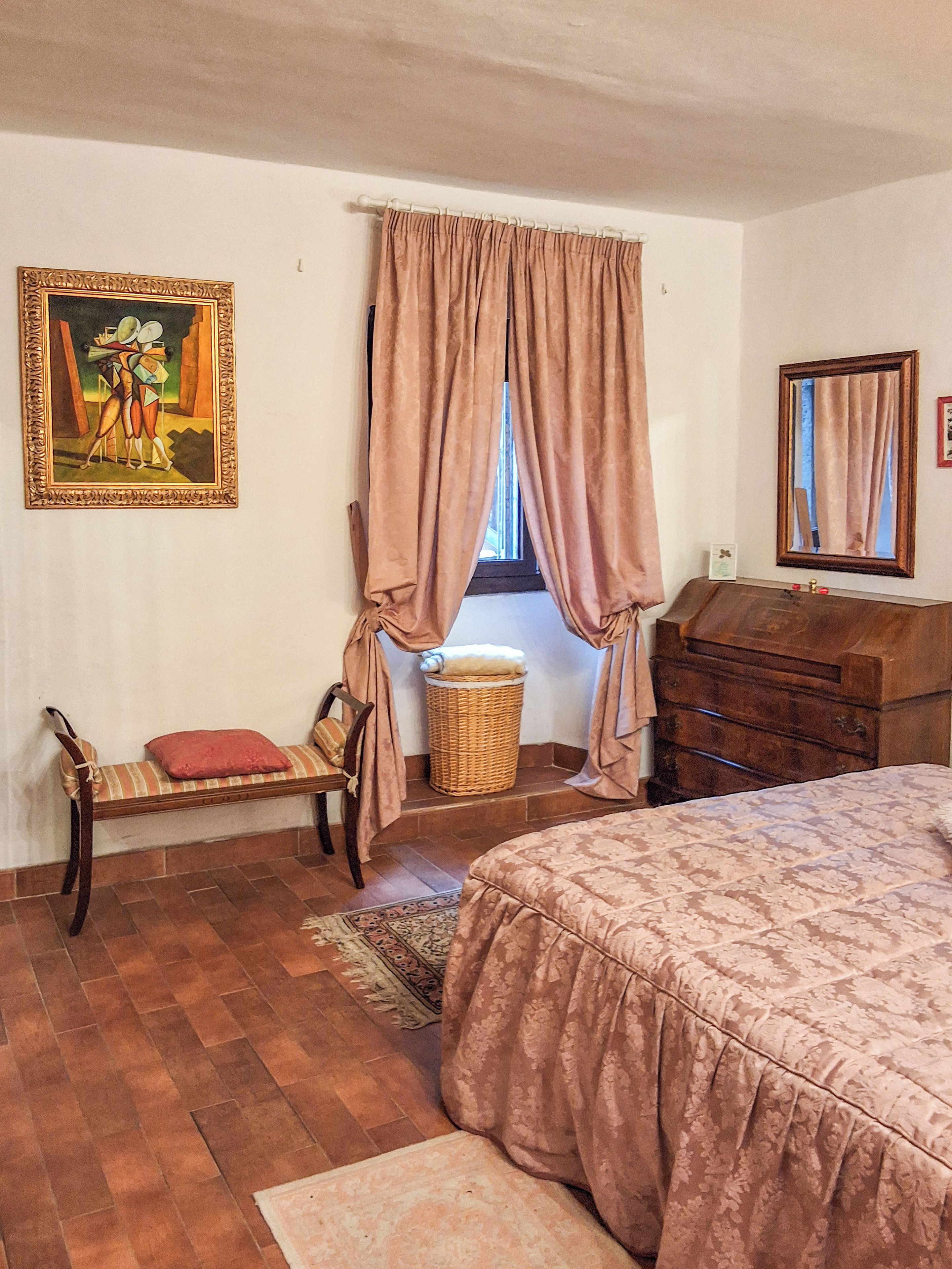A bedroom with a bed, a wooden dresser, a chair, a wicker basket, and a painting on the wall.