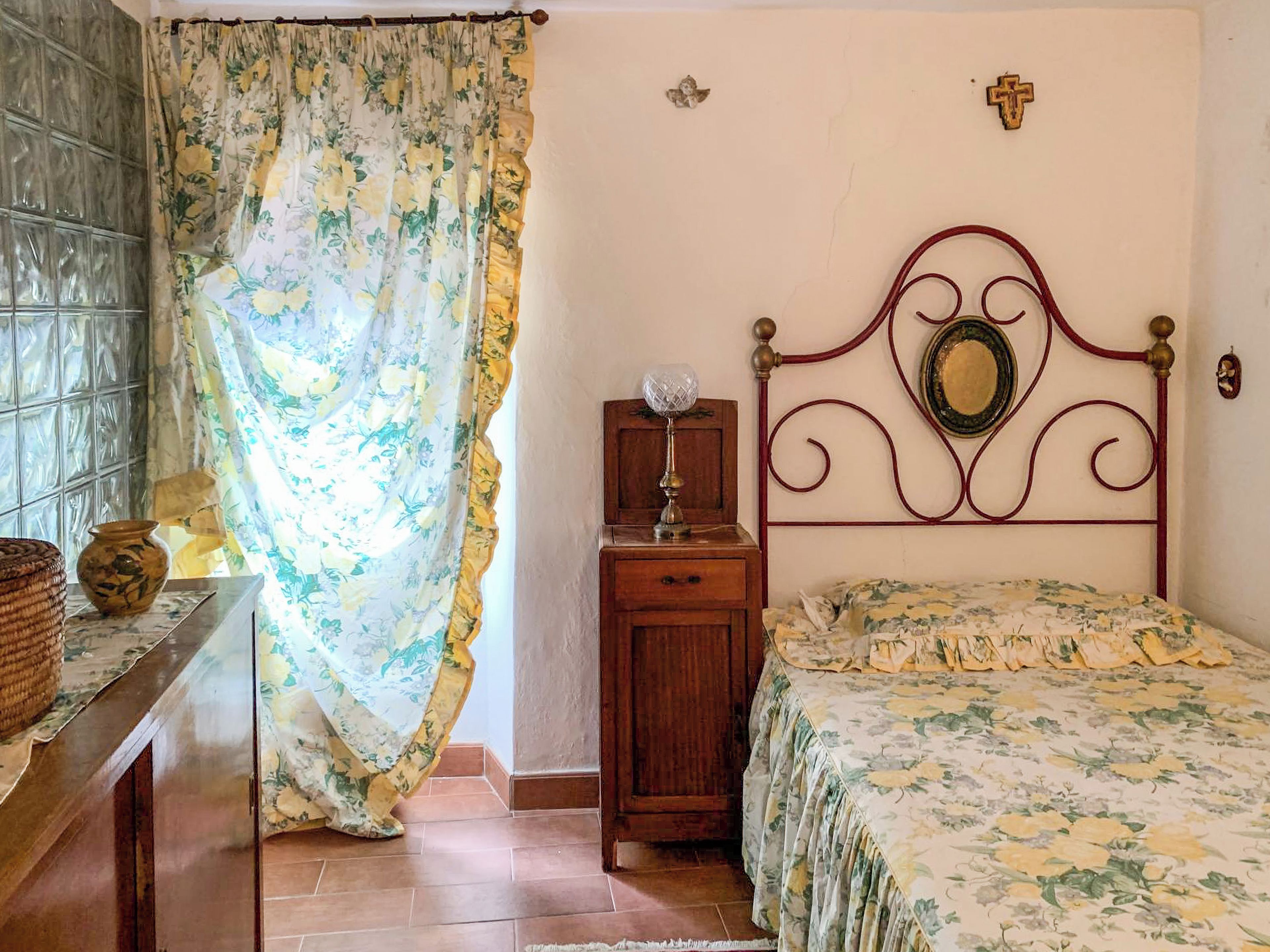 Un dormitorio con cama, mesita de noche, cómoda, ventana con cortinas y una cruz en la pared.