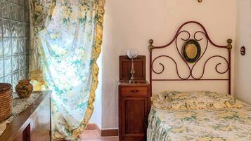 Un dormitorio con cama, mesita de noche, cómoda, ventana con cortinas y una cruz en la pared.