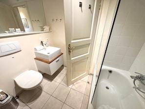 A bathroom with a white sink, toilet, and bathtub. There is a mirror above the sink and a door leading to another room. - Deichhof Apt. 22 (Dunsum)