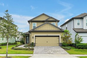 A two-story residential house with a beige garage door, a front yard with a paved driveway, and a small tree. - Magical Moments at Reunion Village Retreat (Kissimmee)