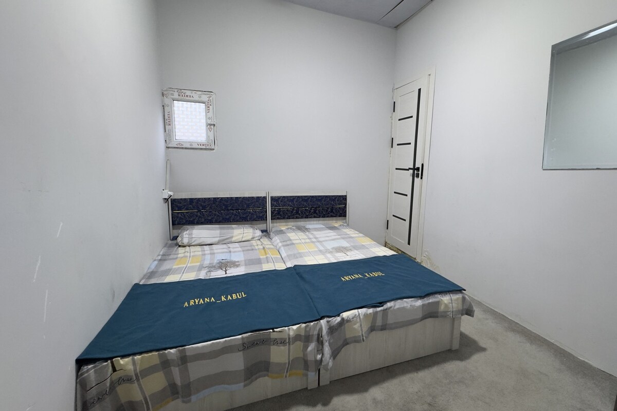 A small, single-bed room with a plain white wall, a window with a grid pattern, a door with a handle, and a mirror on the wall.