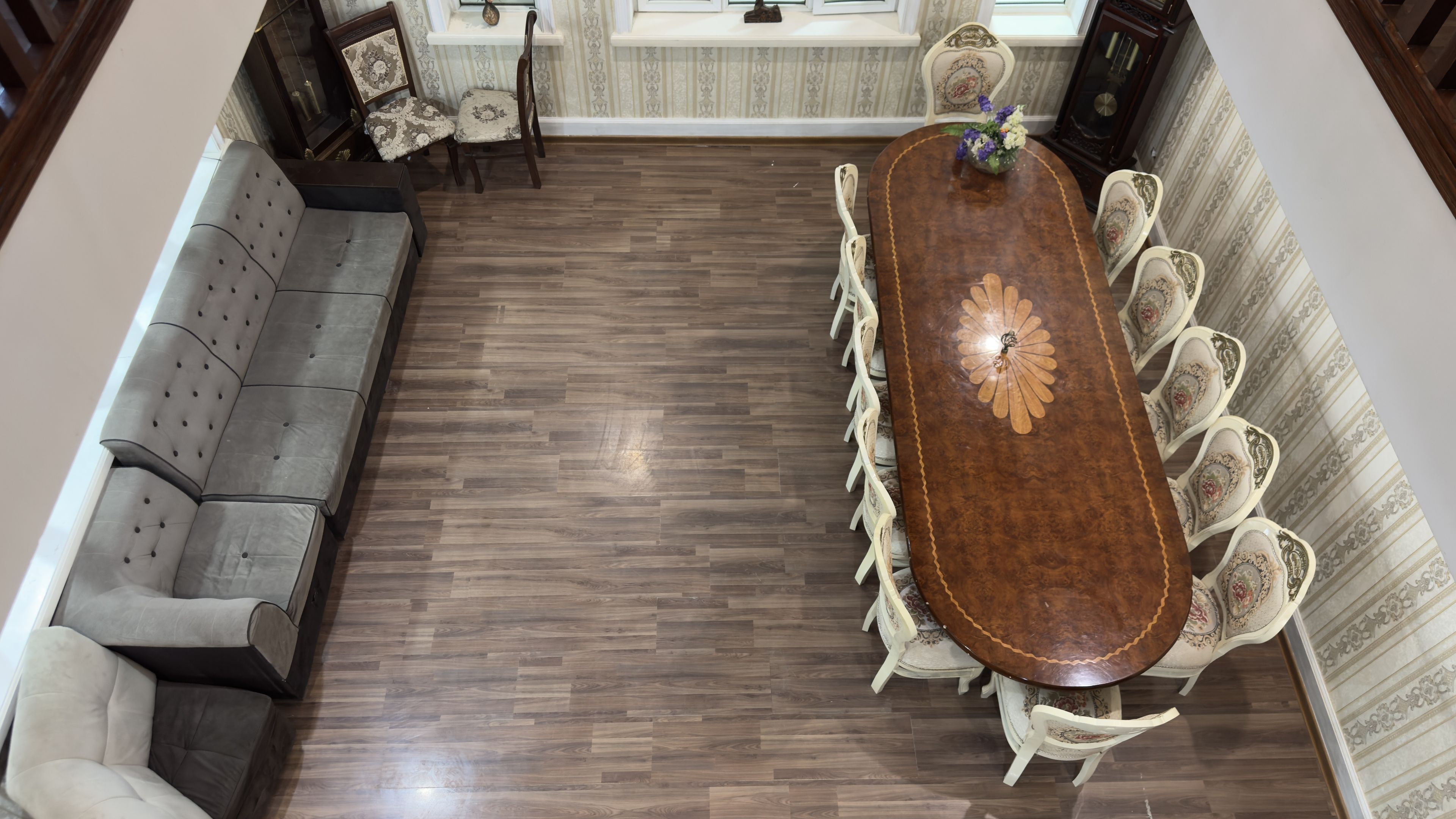 Un comedor con una larga mesa de madera rodeada de sillas blancas con diseños florales, un sofá gris y una mesita con un jarrón de flores.
