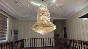 A grand chandelier with multiple tiers of lights, a wooden staircase with balusters, and a patterned wallpaper interior. - ARYANA KABUL HOTEL (Tashkent)