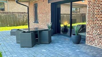 Un espace patio aménagé avec du mobilier d’extérieur, un mur en briques et une porte vitrée donnant sur une entrée abritée.