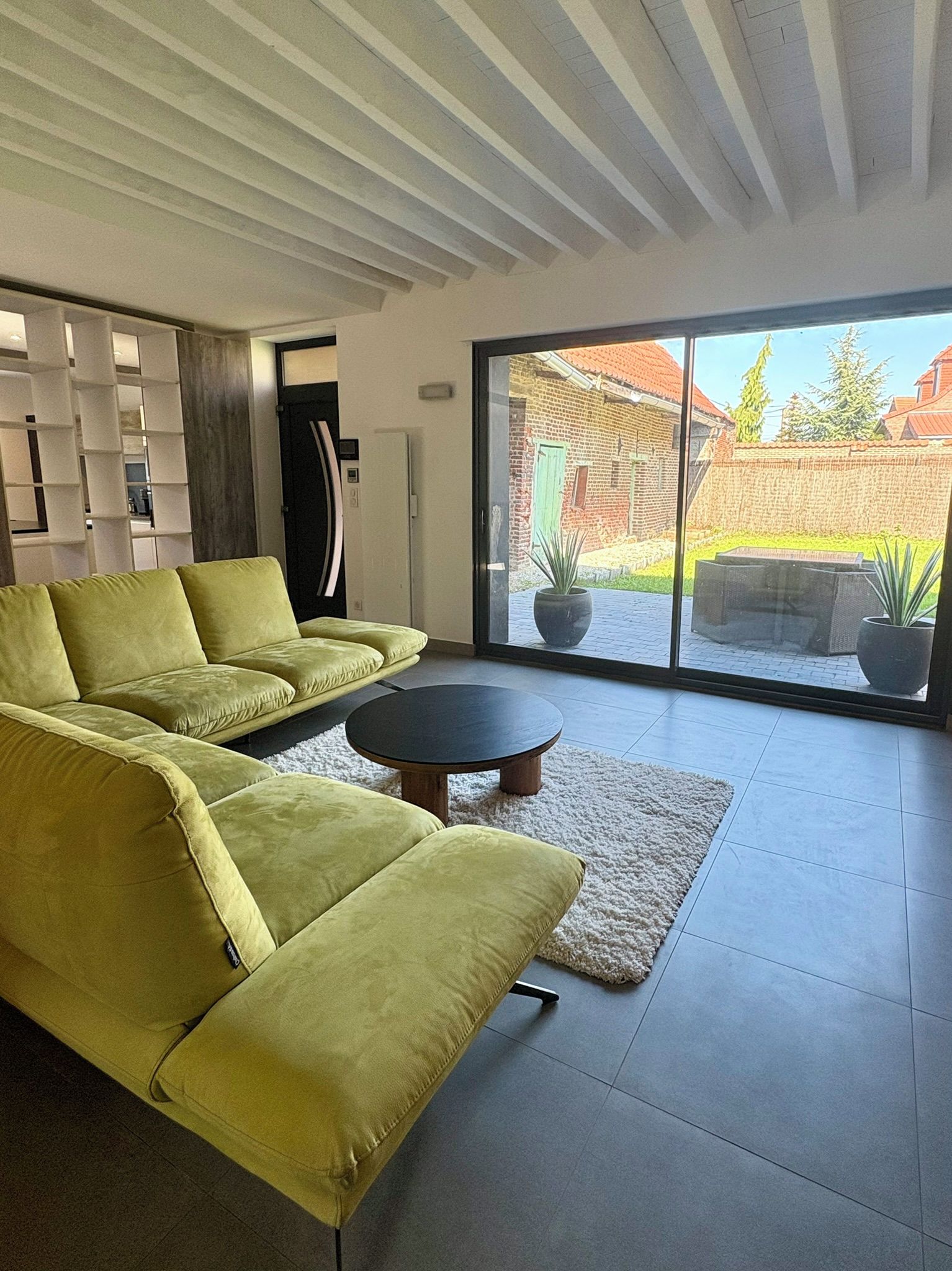 Un salon moderne avec un canapé d’angle, une table basse ronde et une grande porte-fenêtre coulissante donnant sur une terrasse extérieure.