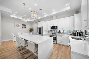 A modern kitchen with white cabinetry, stainless steel appliances, a marble countertop, and a central island with seating. - Refined Coastal 4 Bedroom/4 Bathroom Home with Pool, Spa & Outdoor Kitchen (Naples)