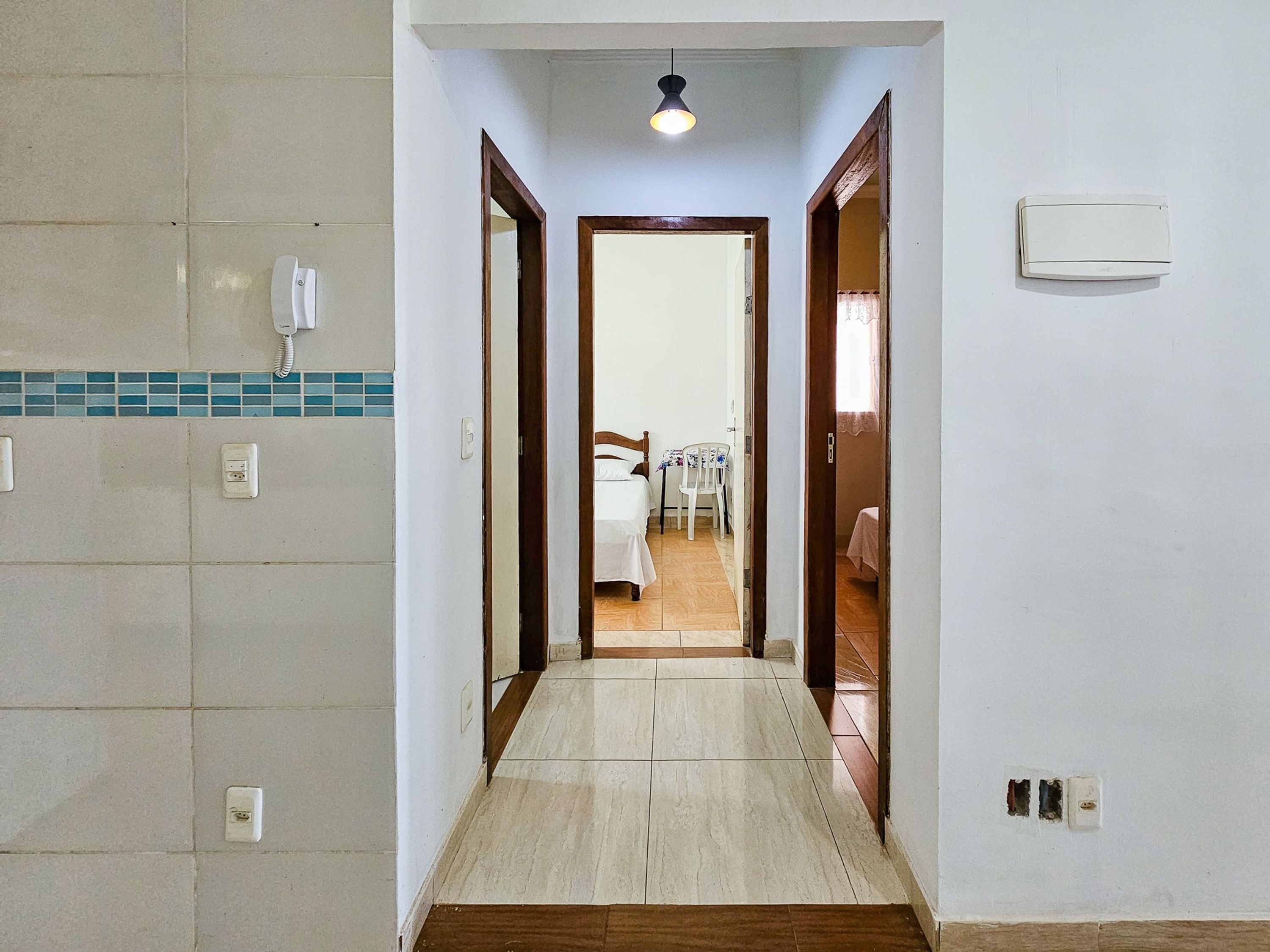 A hallway leading to a bedroom with a bed, a chair, and a table. There are electrical outlets and a light fixture on the walls.