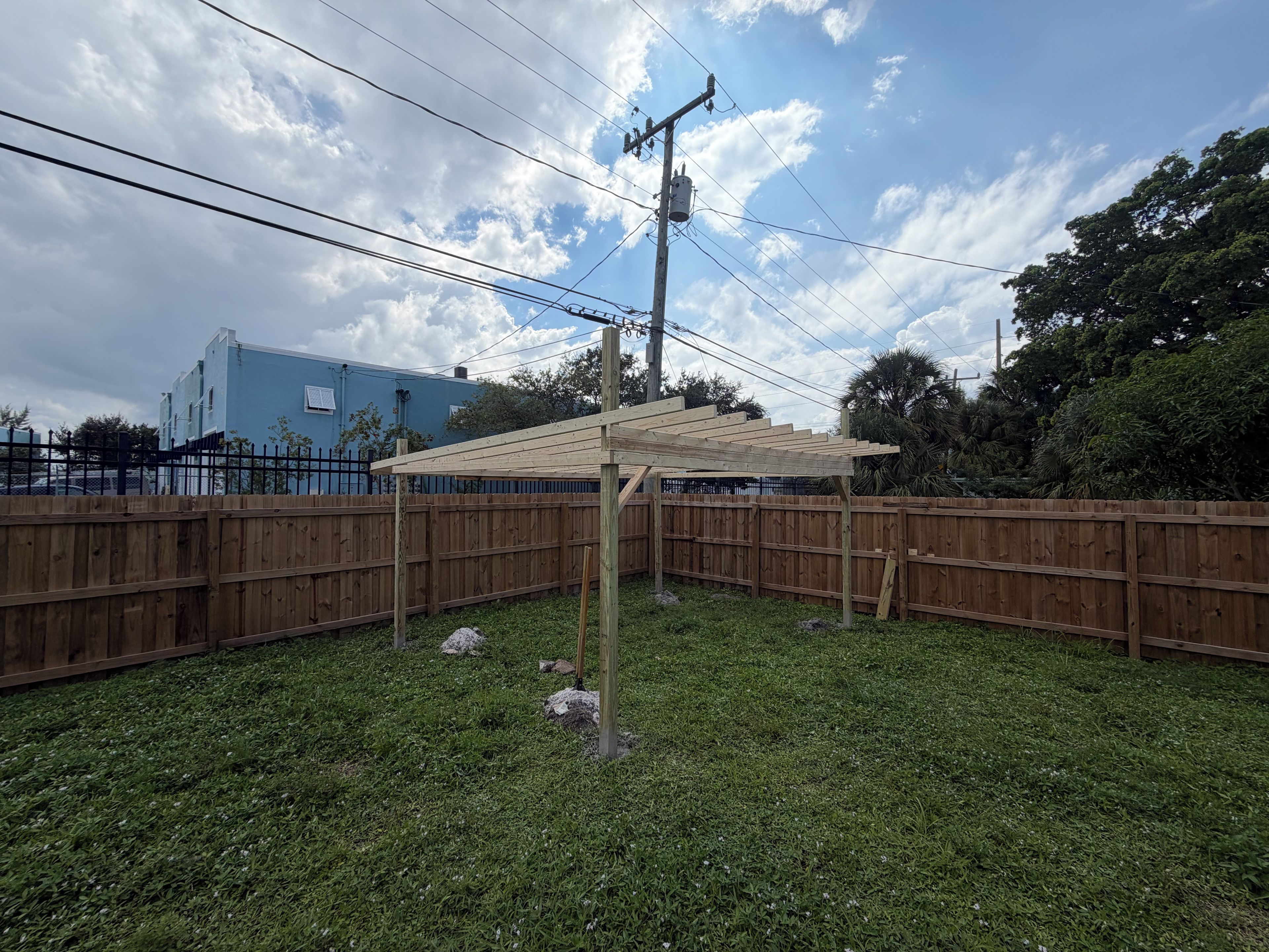 Un patio trasero cercado con una pérgola de madera, un área de césped y un edificio azul al fondo.