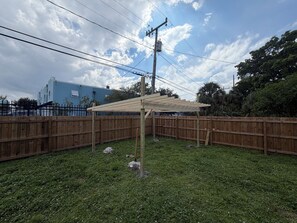 Ein von einer Hecke umgebener Garten mit einer Holz-Pergola, einer Rasenfläche und einem blauen Gebäude im Hintergrund.