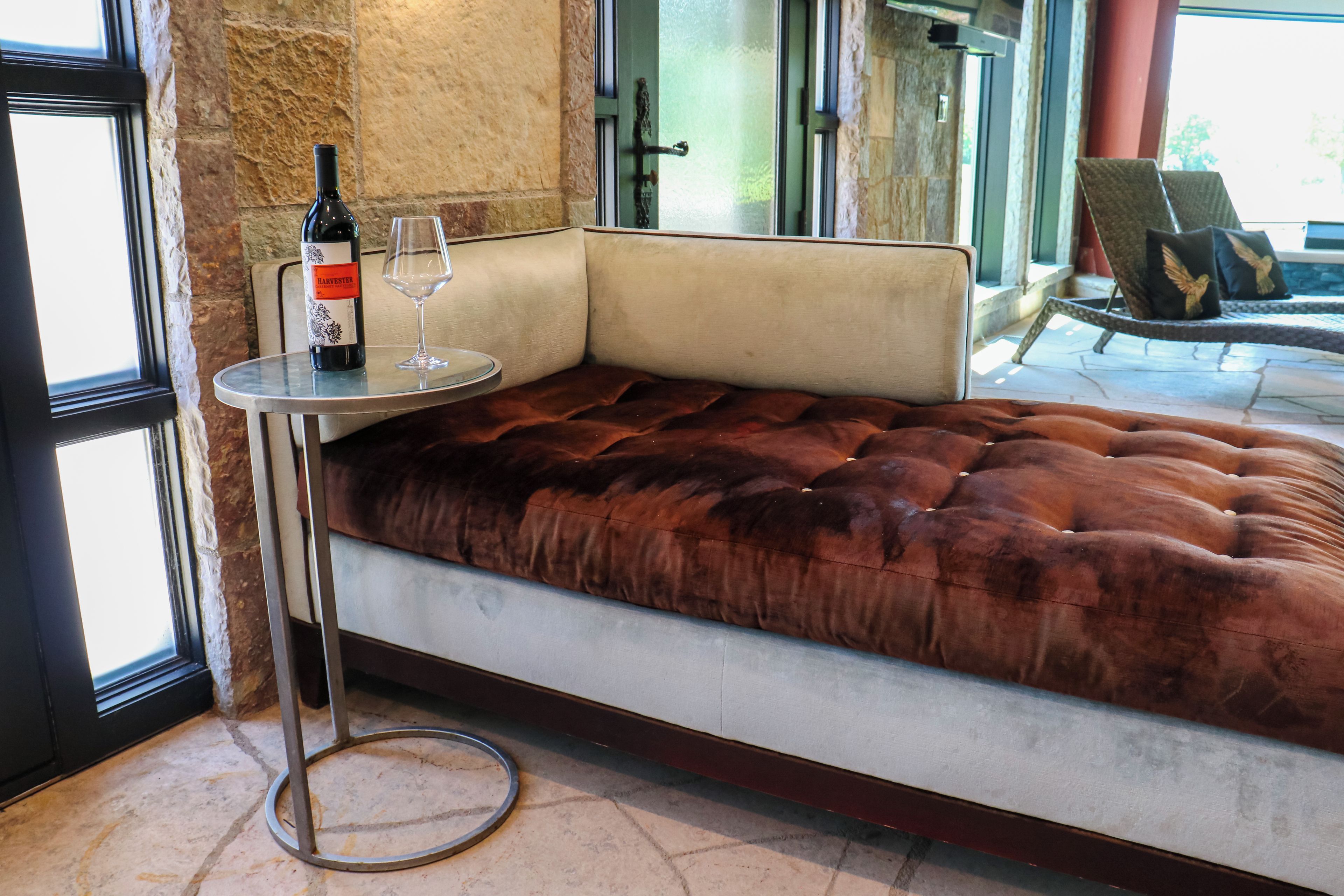 Un rincón con asientos que incluye un sofá de terciopelo capitonado, una mesa de vidrio con una botella de vino y una copa, y una pared de piedra al fondo.