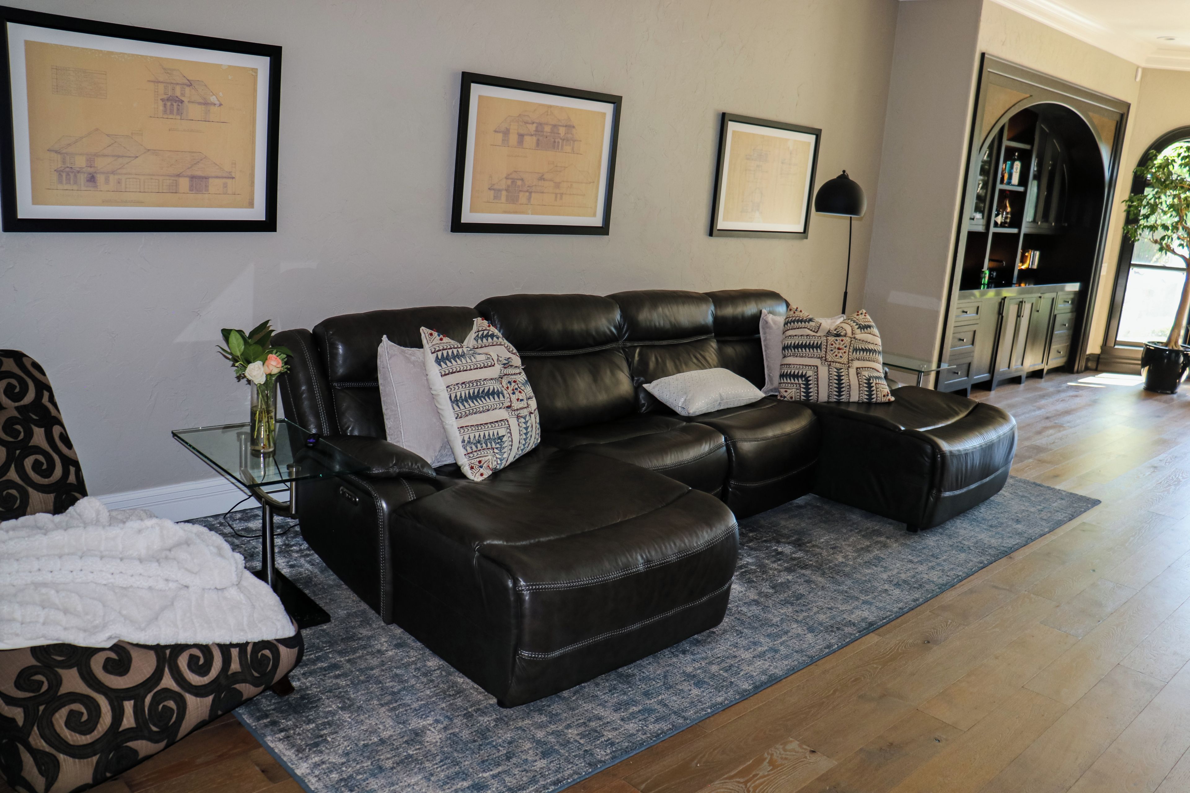 A living room with a leather sectional sofa, a glass coffee table, and a patterned rug. There are framed architectural drawings on the wall.