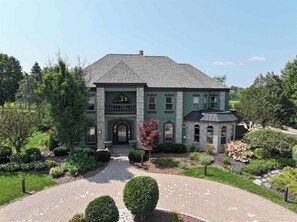 A two-story house with a brick facade, a central entrance with a decorative arch, and a landscaped garden with various shrubs and a paved driveway. - Indoor Pool-Golf Simulator-RaceCar Simulator-HotTub-Movie Theatre-PoolTables (Fond du Lac)