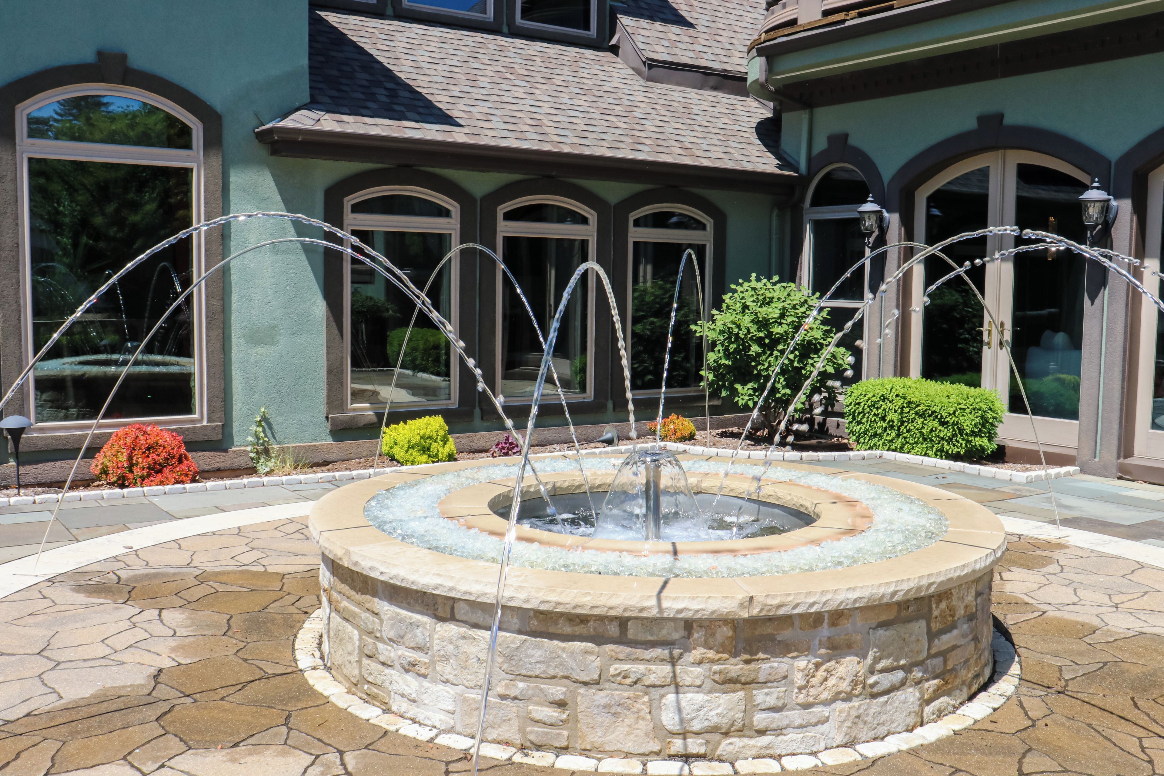 A stone fountain with water jets in a circular basin, surrounded by a stone pathway and a building with arched windows and a tiled roof.