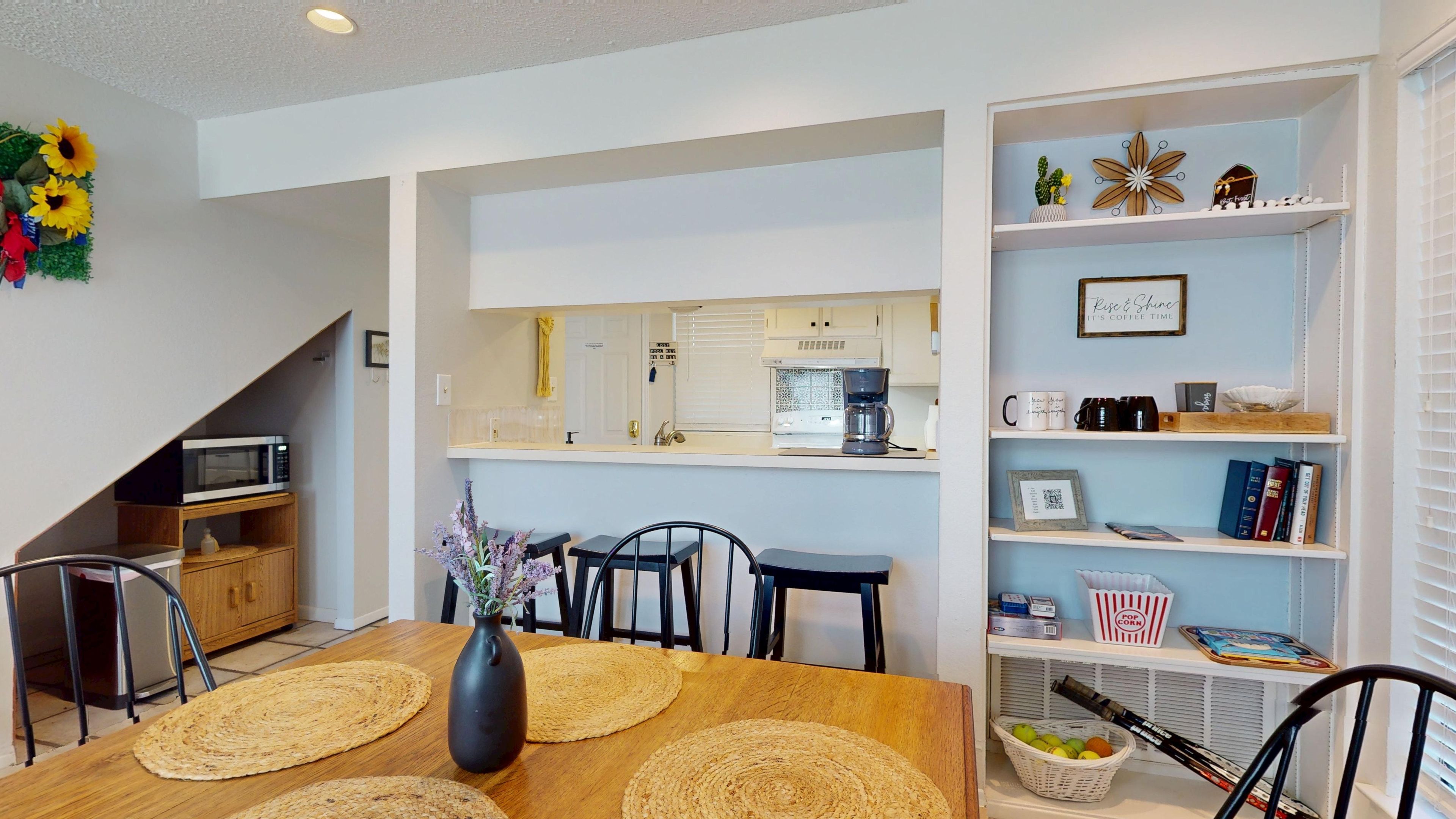 A modern kitchen with a microwave, coffee maker, and a small refrigerator. There is a dining area with a wooden table, chairs, and a vase with flowers. A bookshelf with various items is visible in the background.