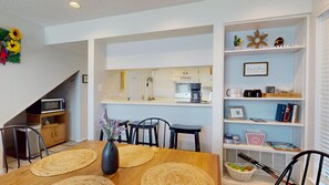 A modern kitchen with a microwave, coffee maker, and a small refrigerator. There is a dining area with a wooden table, chairs, and a vase with flowers. A bookshelf with various items is visible in the background. - River City Resorts: Among the Oaks T-Bar-M 112: Spacious Hill Country Condo. (New Braunfels)