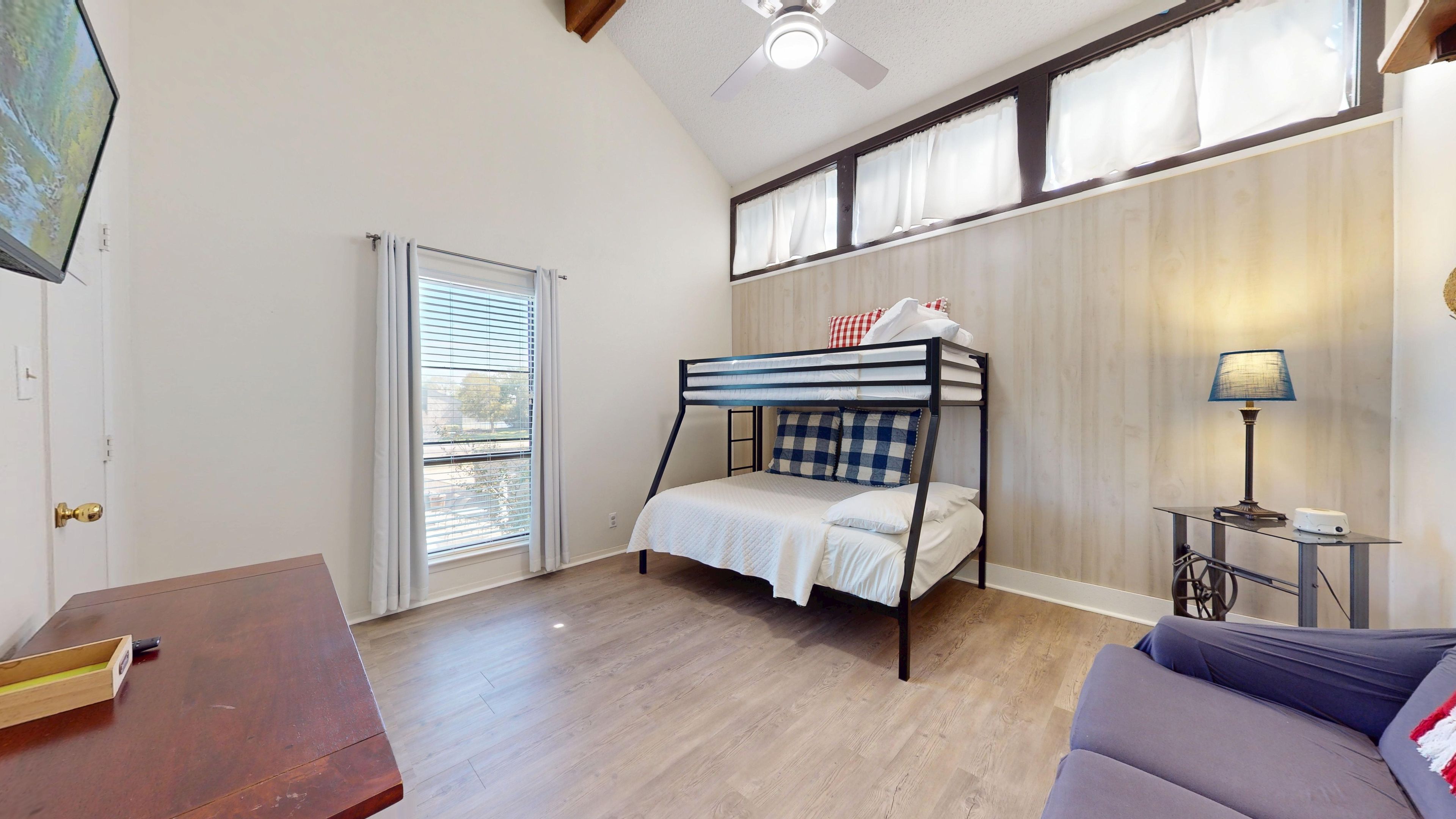 A room with a bunk bed, a desk, a lamp, and a window with blinds.