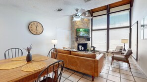 A living room with a sofa, a dining table with placemats, a clock, and a fireplace. - River City Resorts: Among the Oaks T-Bar-M 112: Spacious Hill Country Condo. (New Braunfels)