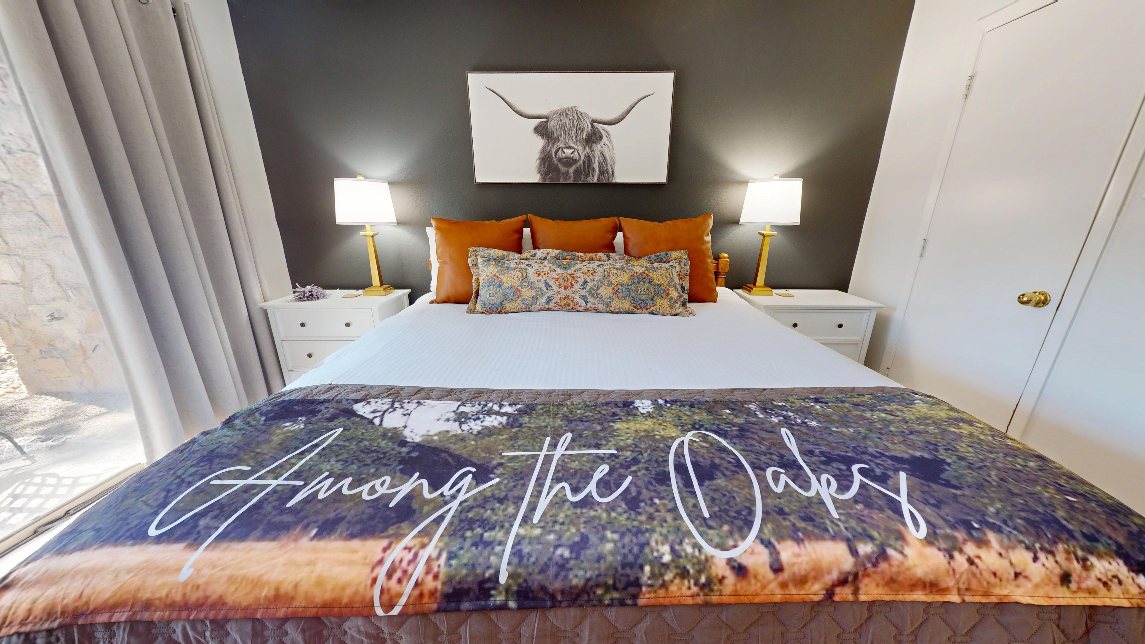A hotel room with a bed, two bedside lamps, a framed artwork above the bed, and a window with curtains.