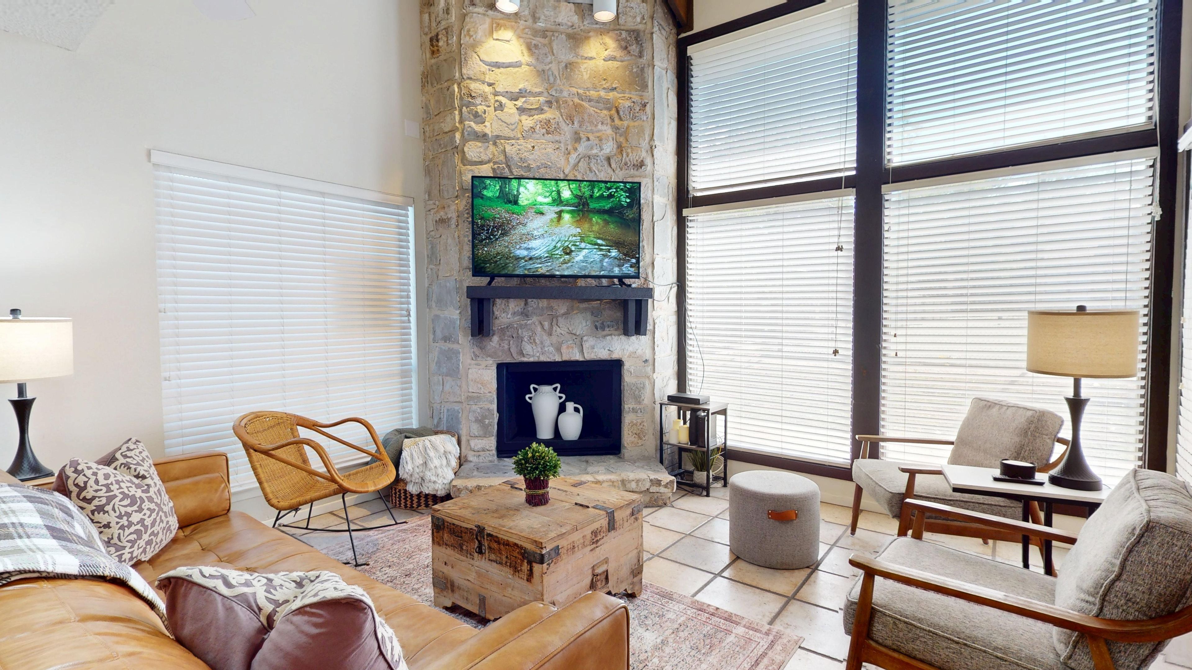 A cozy living room with a stone fireplace, comfortable seating, and large windows.