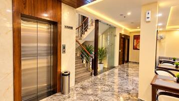 A hotel lobby with a marble floor, an elevator, a staircase, and a seating area with tables and chairs.
