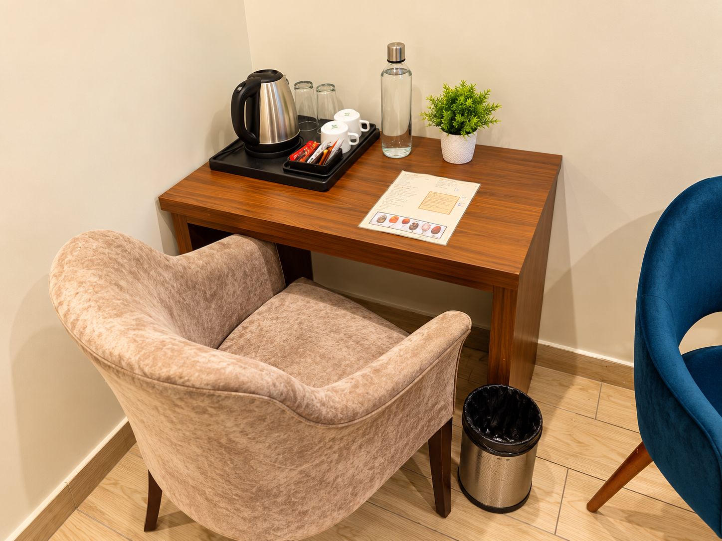 Un pequeño escritorio de madera con una tetera, tazas de café, una botella y un menú. Hay un cubo de basura y una silla con cojín.