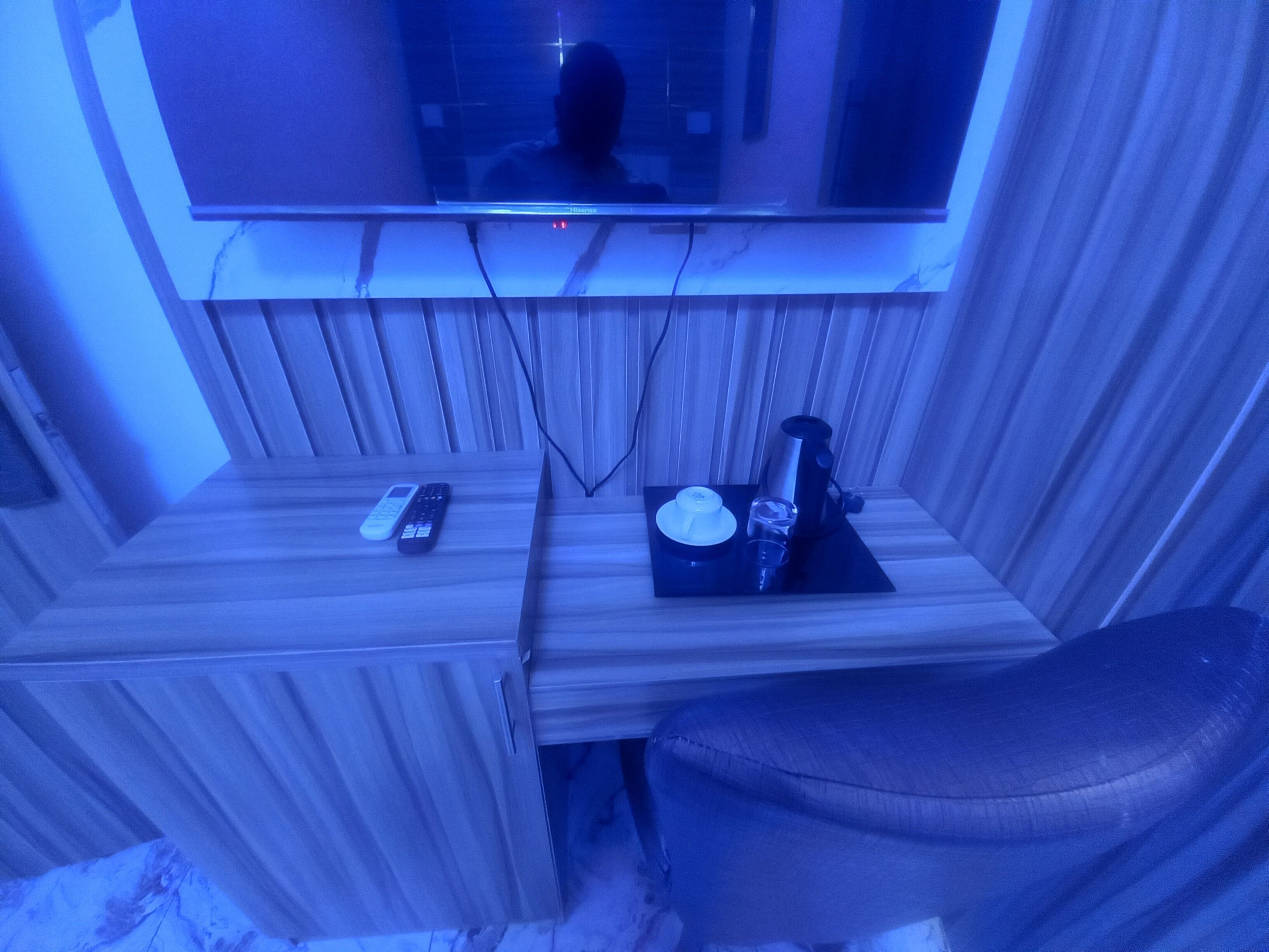 Una habitación con un televisor montado en una pared de paneles de madera, un control remoto, una taza y un vaso sobre una pequeña mesa de madera.