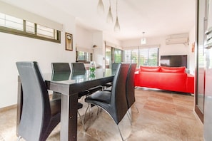 A dining area with a glass table and black chairs, a red sofa in the background, and a TV mounted on the wall.