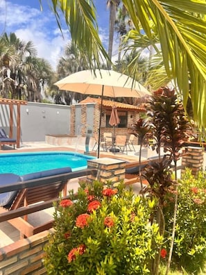 Un área de piscina con una piscina azul cristalina, rodeada de palmeras y una estructura de ladrillo.