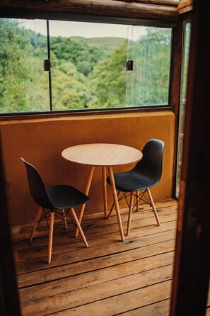 Eine Holzterrasse mit einem runden Tisch und zwei Stühlen, mit Blick auf eine bewaldete Landschaft durch ein großes Fenster.