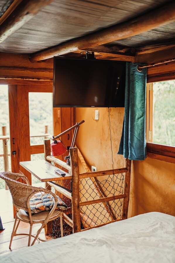 Ein gemütliches Cabin-Interieur mit einem Bett, einem an der Wand montierten Fernseher, einem Holztisch mit einer roten Stricktasche und einem Korbstuhl.