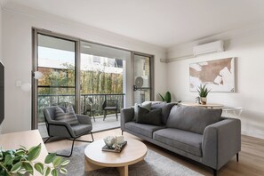 A modern living room with a grey sofa, a round coffee table, and a grey armchair. There is a sliding glass door leading to an outdoor balcony with a view of bamboo and a brick wall. - Urban Rest Bondi Park (Bondi)