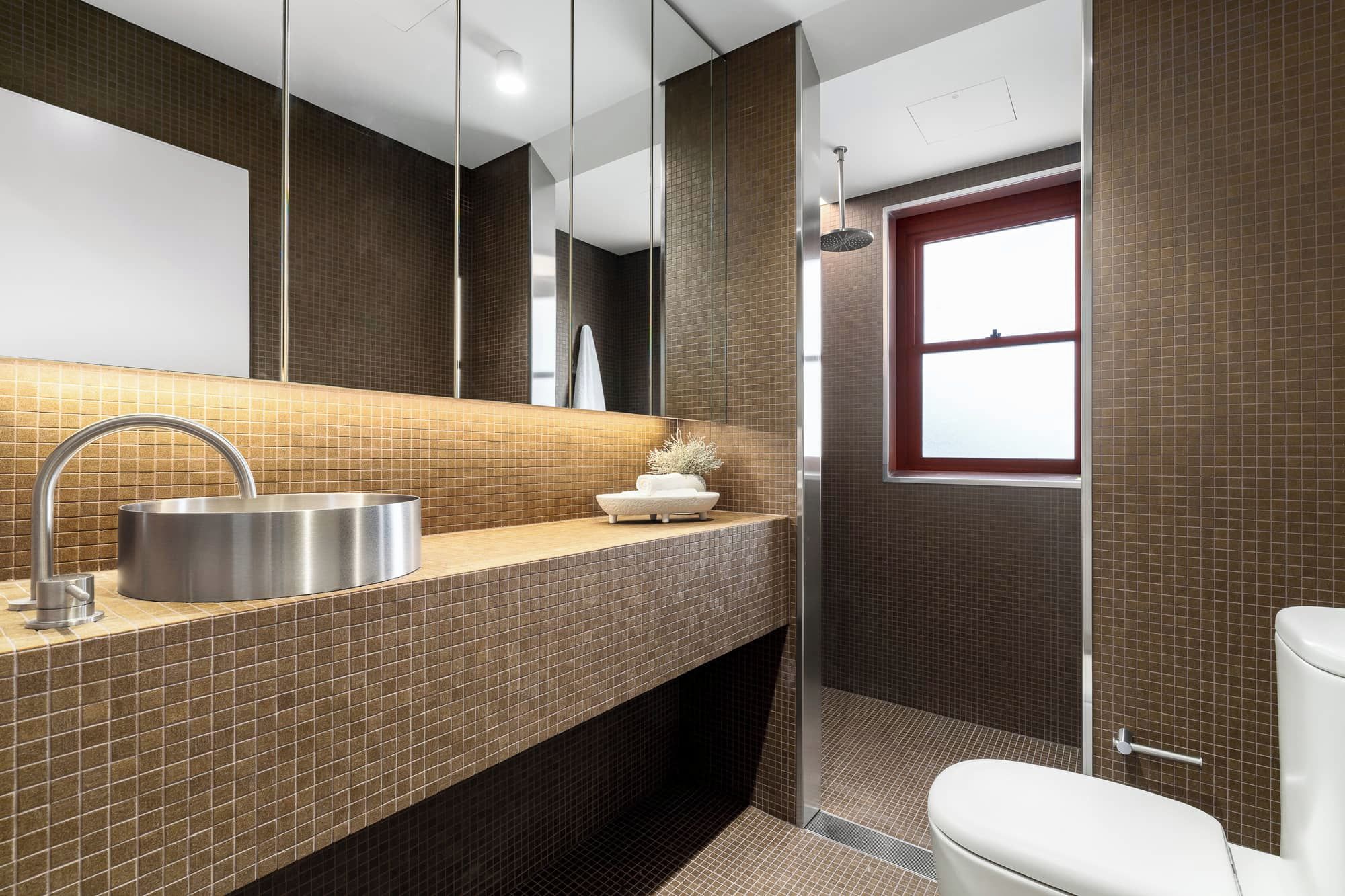 Un baño moderno con un espejo grande, un lavamanos de acero inoxidable, un inodoro y una zona de ducha.