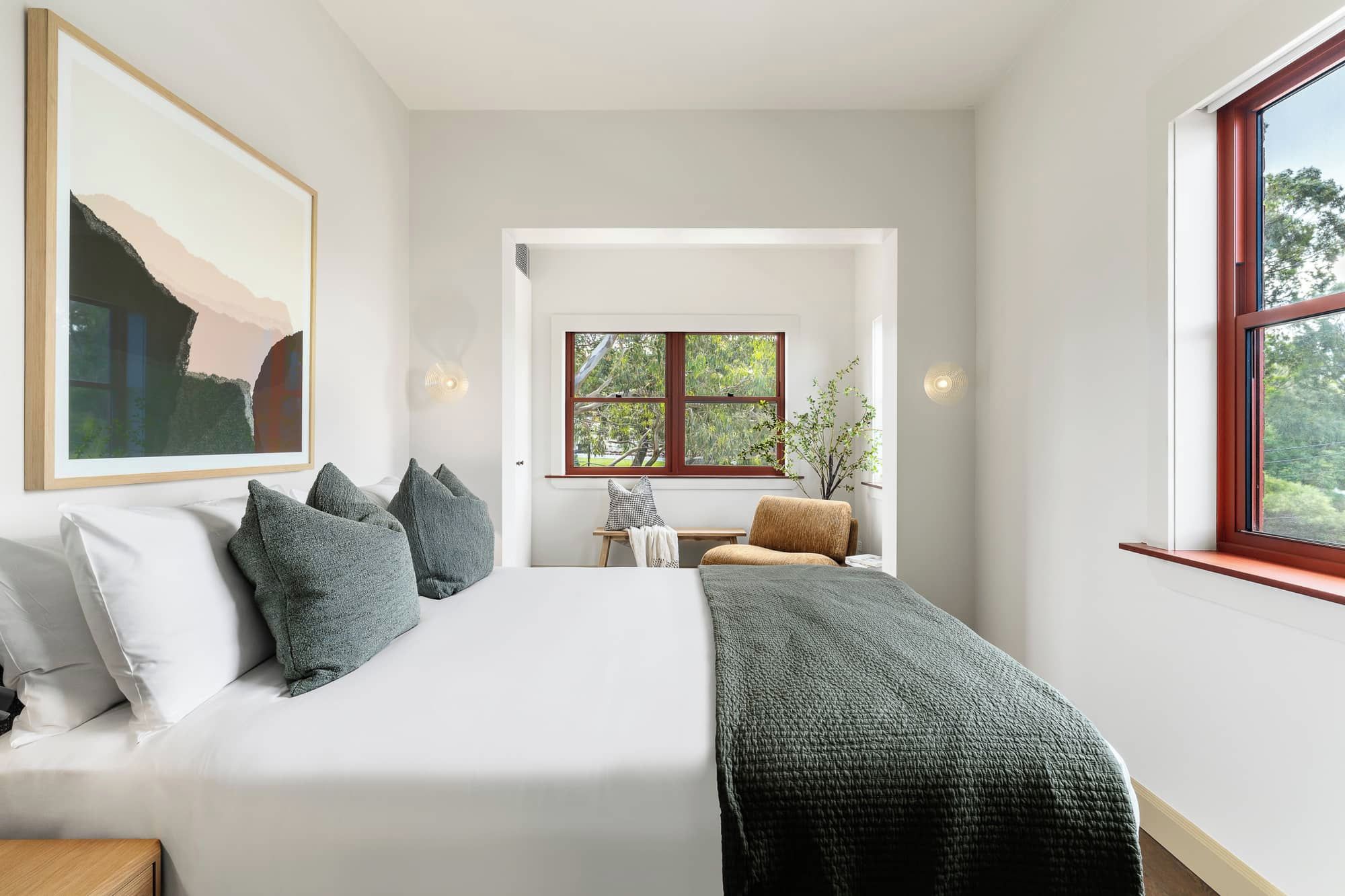 Un dormitorio con cama, almohadas, una mesita de noche, una silla, una ventana y un cuadro en la pared.