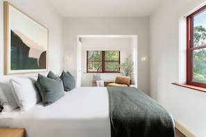 Un dormitorio con cama, almohadas, una mesita de noche, una silla, una ventana y un cuadro en la pared.