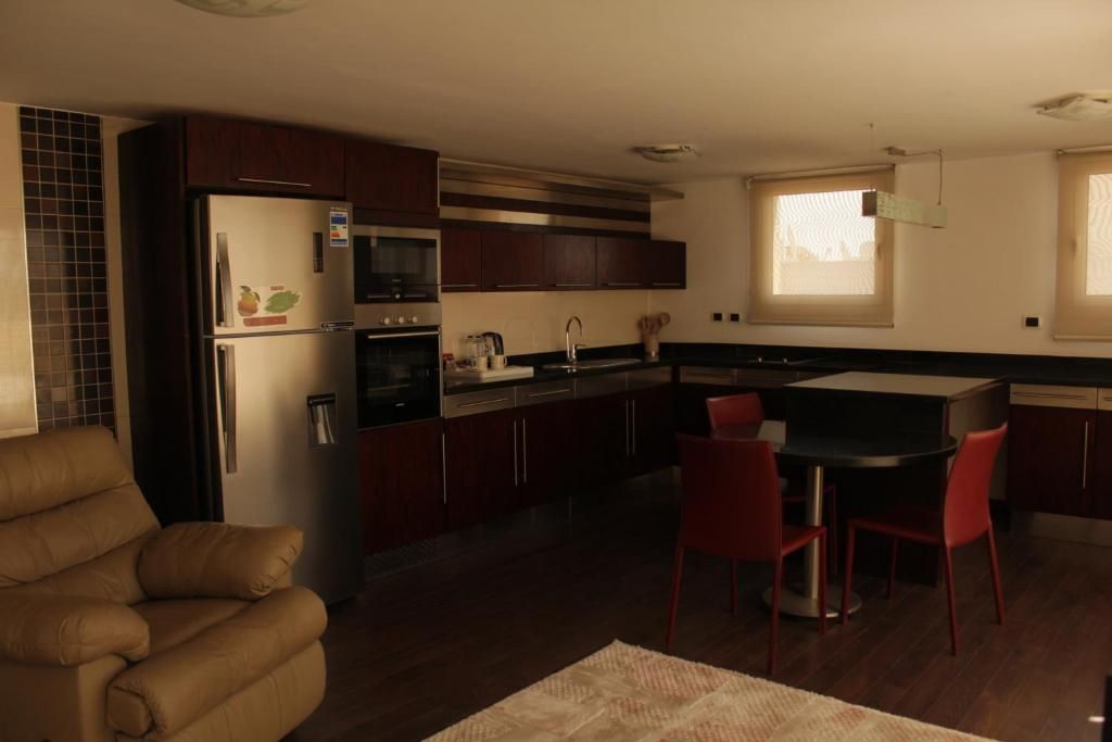 Cocina compacta con alacenas de madera oscura, refrigerador, microondas y una zona de comedor con sillas rojas.
