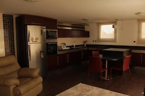 A compact kitchen with dark wooden cabinets, a refrigerator, microwave, and a dining area with red chairs. - The 88th Signature by Maran Hospitality (New Cairo)