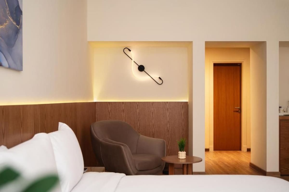 A hotel room with a bed, a chair, a small table with a plant, and a wall-mounted light fixture.
