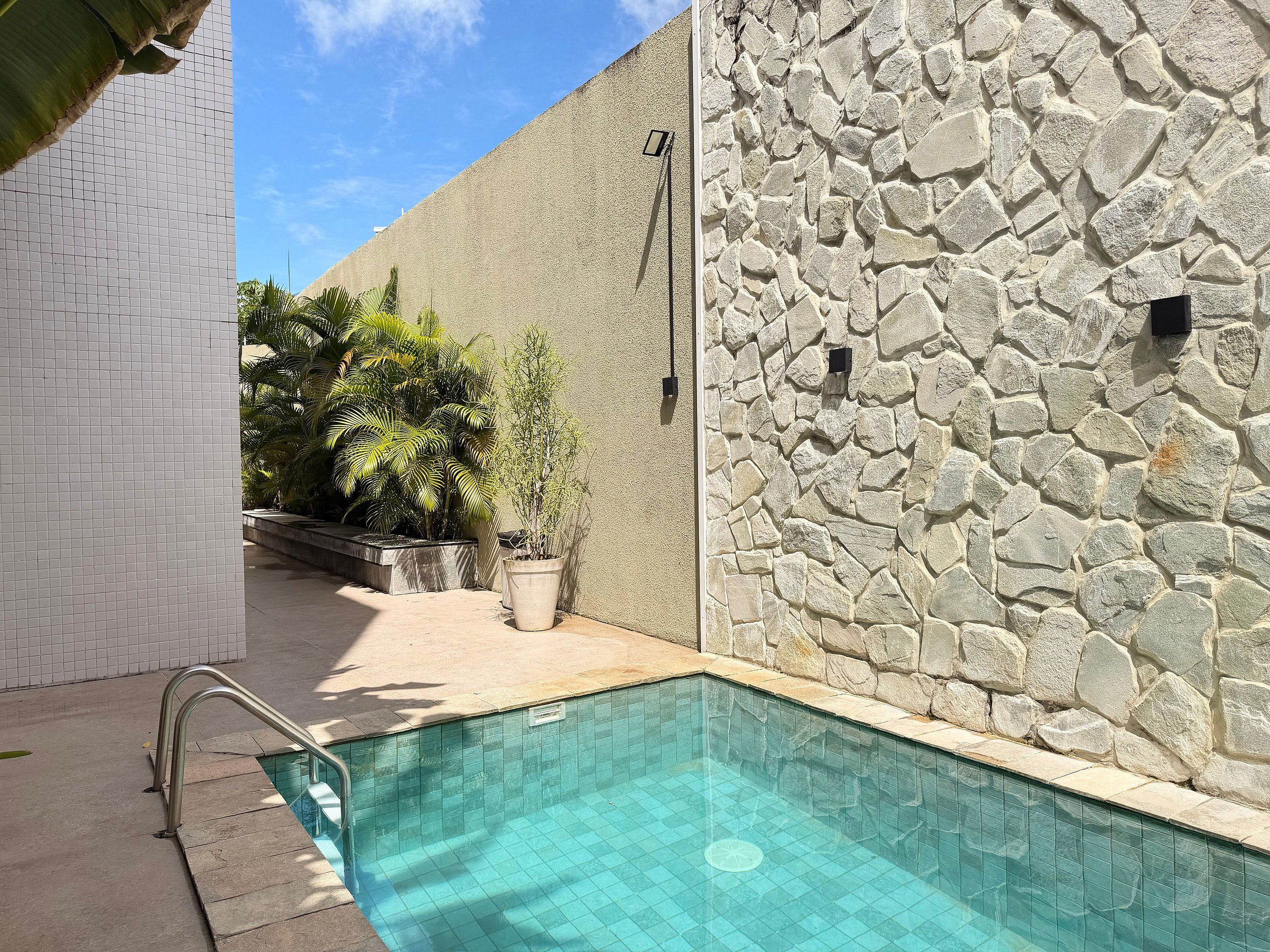 Una piscina con pared de piedra y suelo de baldosas, rodeada de plantas en macetas y una pared de baldosas blancas.