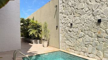 Una piscina con pared de piedra y suelo de baldosas, rodeada de plantas en macetas y una pared de baldosas blancas.