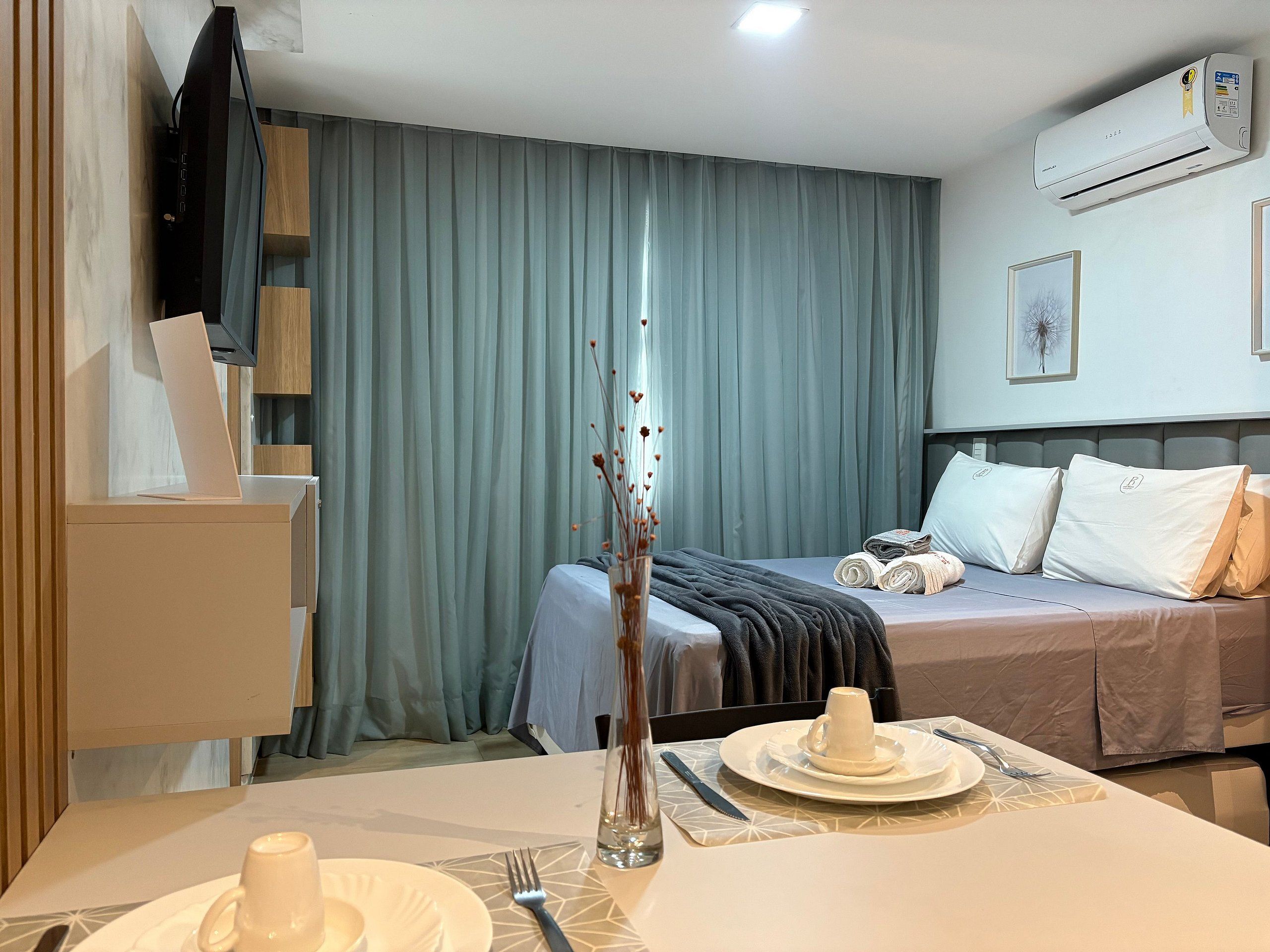 A hotel room with a bed, a desk with a laptop, a dining table set for two, and an air conditioning unit on the wall.