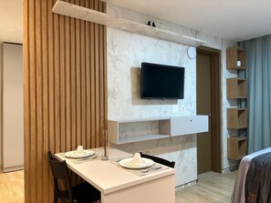 Un comedor moderno con una mesa blanca, dos sillas y un televisor de pantalla plana montado en una pared con textura similar al mármol.