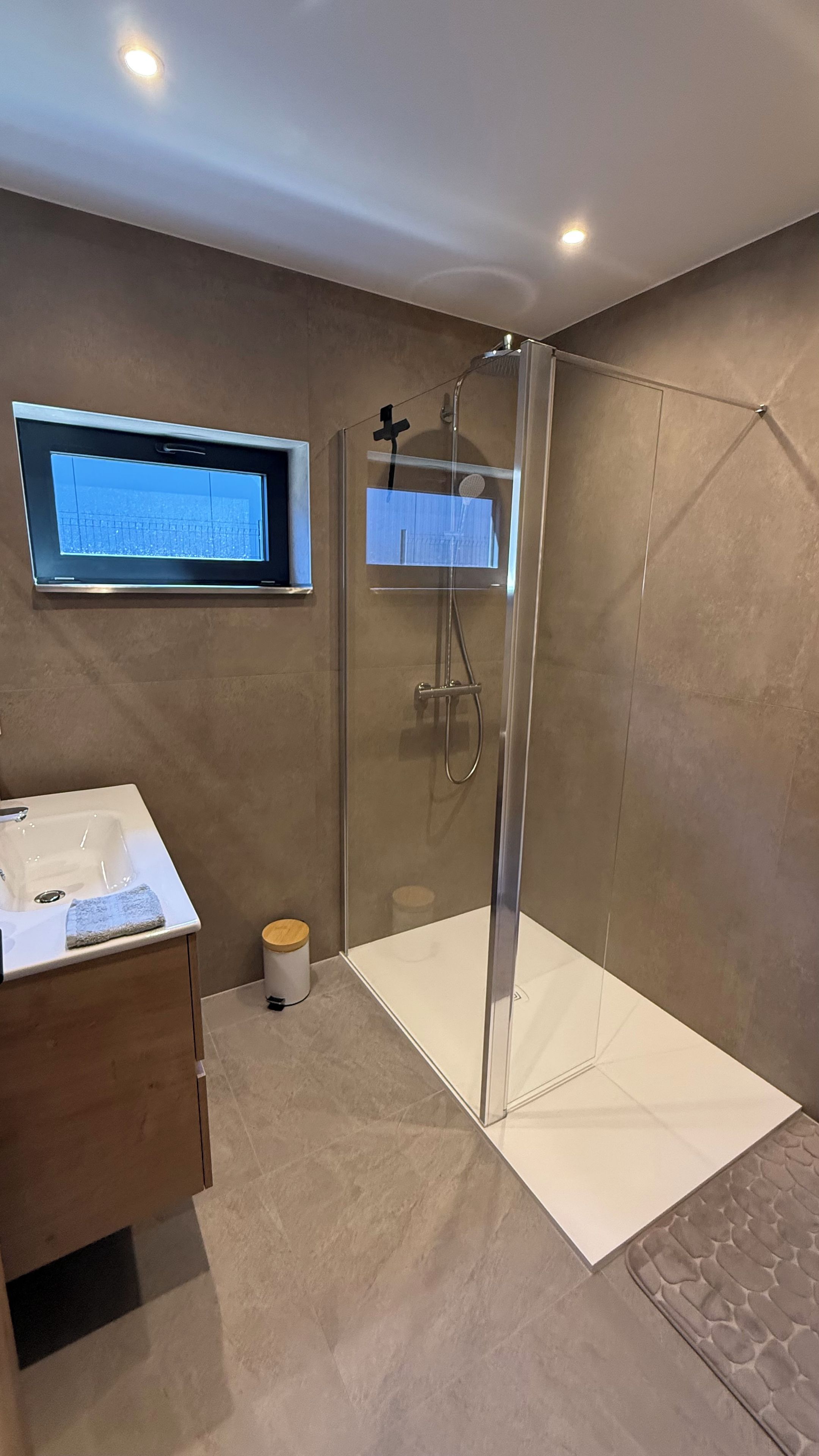 Een moderne badkamer met een douchecabine van glas, een wastafelmeubel met een wastafel, en een raam boven de spiegel.