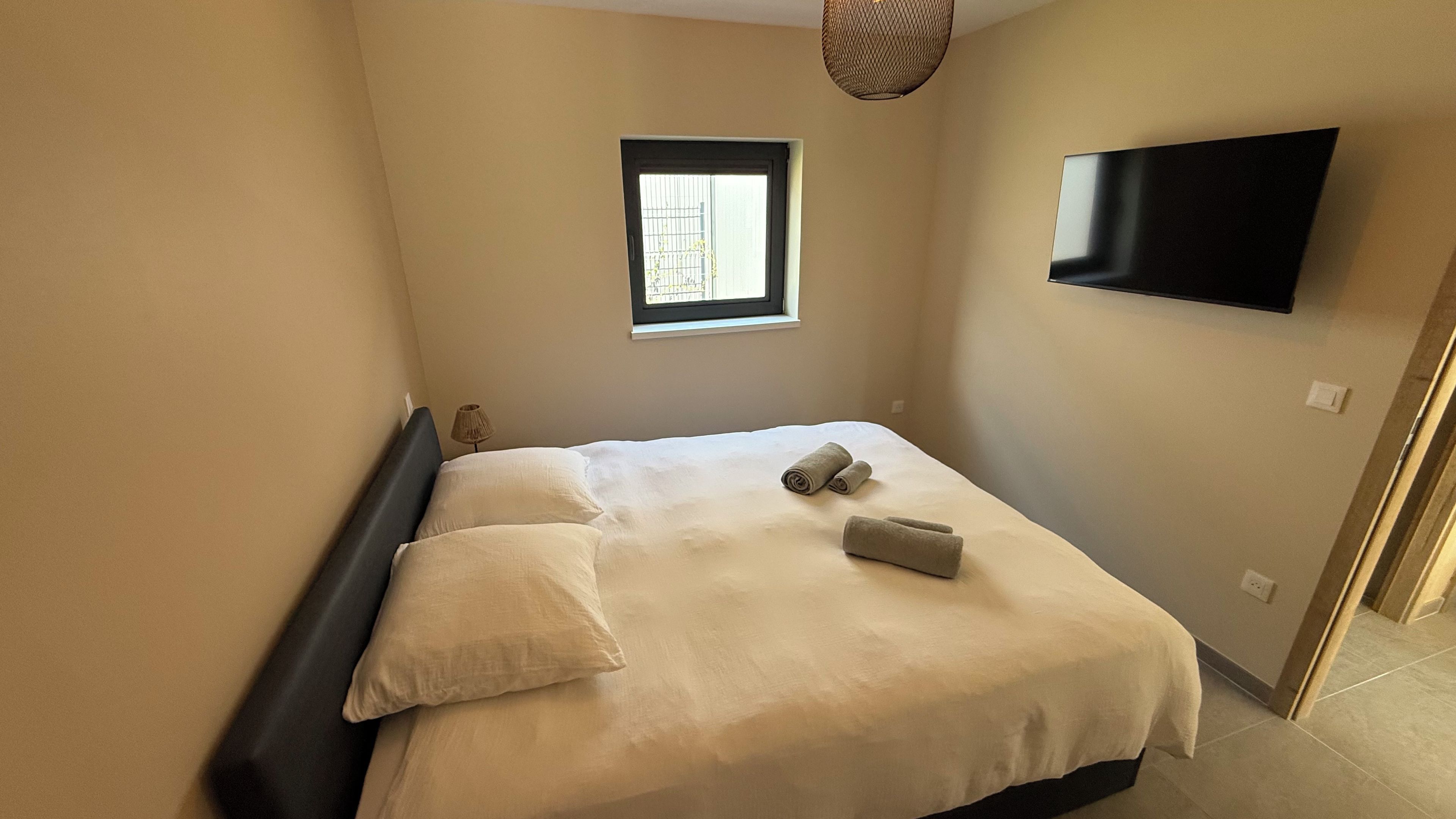 Een slaapkamer met een bed, een raam, een flatscreen televisie en een lamp.