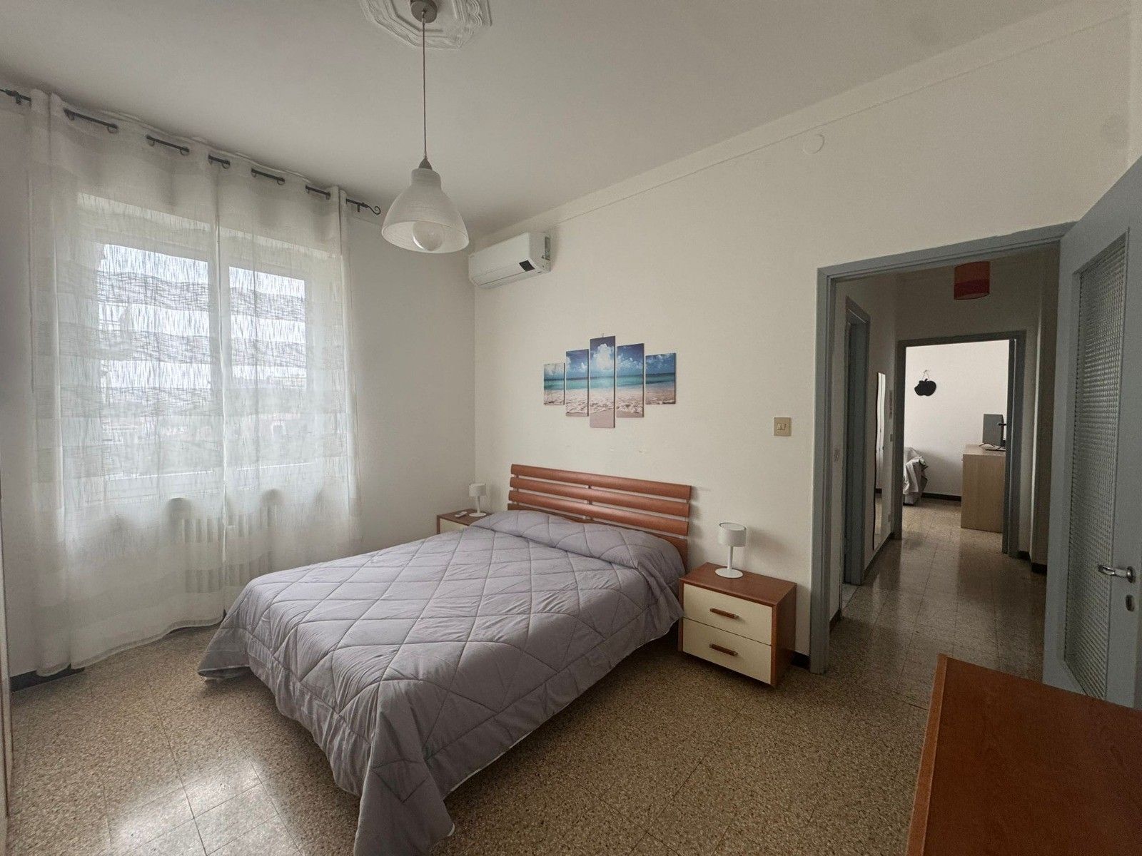 Un dormitorio con cama, mesitas de noche, lámpara colgante, aire acondicionado instalado en la pared y una puerta que da a otra habitación.