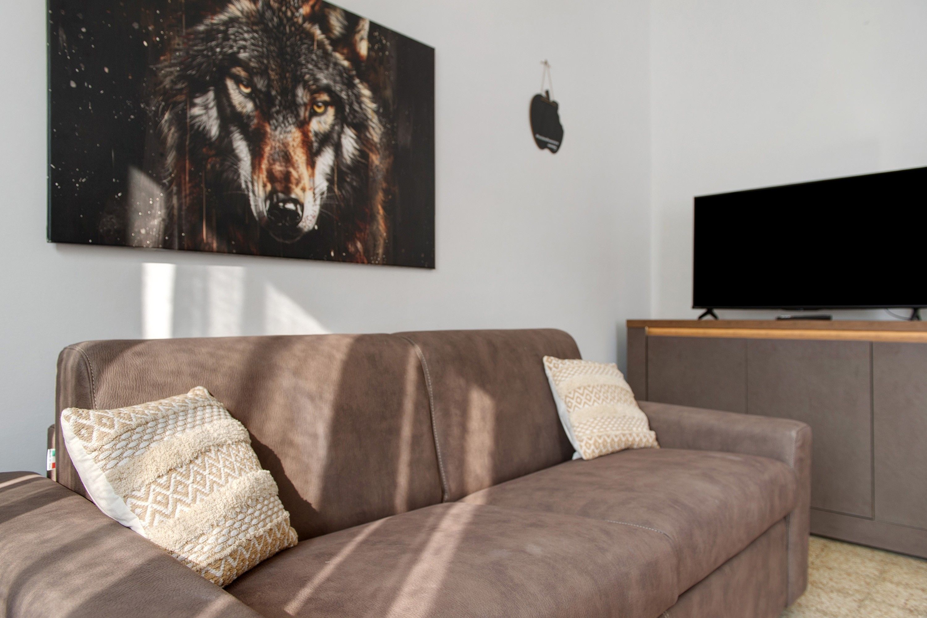 A living room with a brown sofa, two patterned cushions, a large wolf painting on the wall, and a flat-screen TV on a stand.