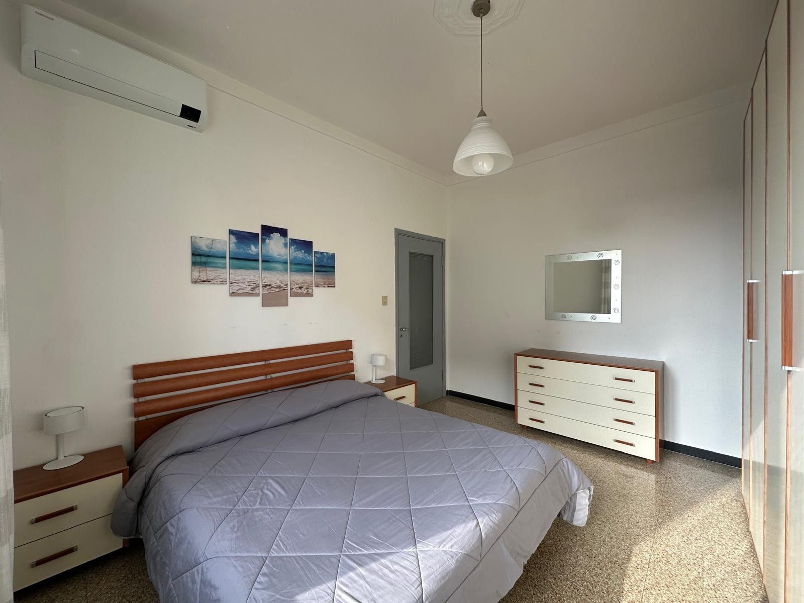 Ein Schlafzimmer mit Bett, Nachttisch, Lampe, Kommode, Spiegel und Wandbildern.